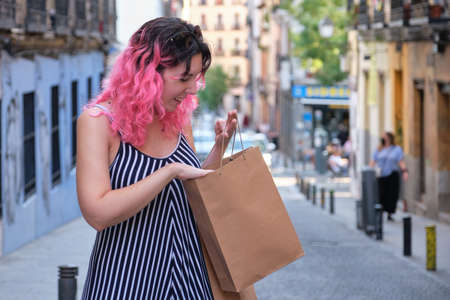 Young caucasian woman with pink hair looking inside of shopping bag in street.の写真素材