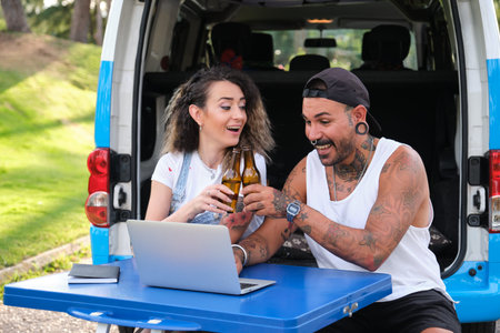 Young tattooed couple smile, make a toast with beer and use the laptop.