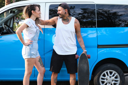 Young tattooed couple smile and look each other near by van with a longboard.