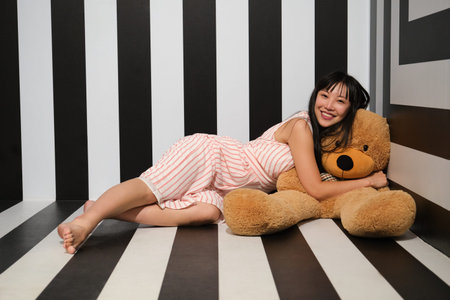 Young asian woman hug and lay over a teddy bear on the striped room floor.