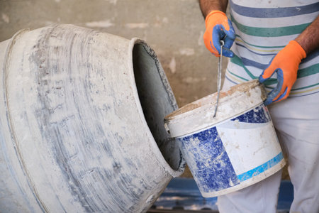 Construction worker with a bucket in his hands loads a concrete mixer.