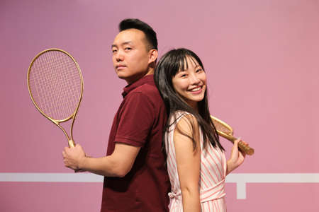 Young asian couple holding tennis racket while posing against pastel pink court.の写真素材