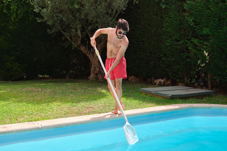 Young man cleaning swimming pool of fall leaves with cleaning net.