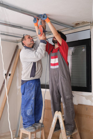 Two workers assembling a suspended ceiling metal frame with a screwdriver.
