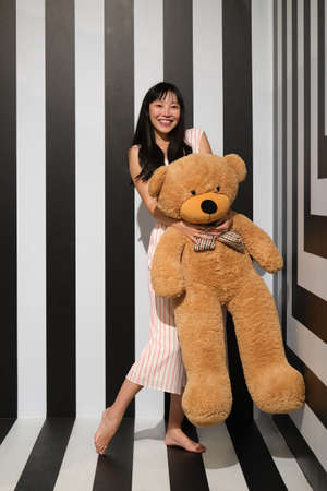 Young asian woman hugging a teddy bear standing on a striped room floor.の写真素材