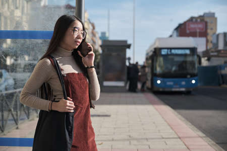 Young asian woman talking on her phone while waiting for the bus at bus stop.の写真素材