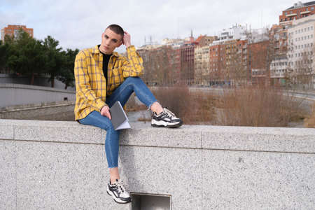 Young non binary person sitting on a wall with a book looking at camera.の写真素材