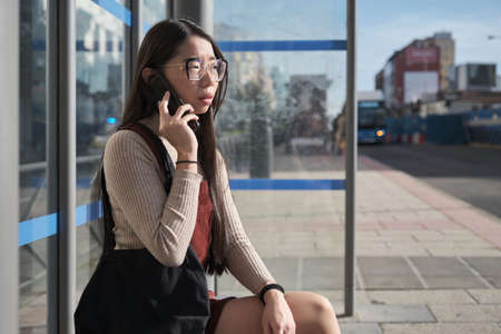 Young asian woman speaking on her smartphone while waiting for the bus at bus stop.の写真素材