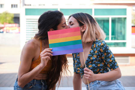 Young lesbian couple kissing and hiding behind a rainbow flag outdoors. LGBT relationship.の写真素材
