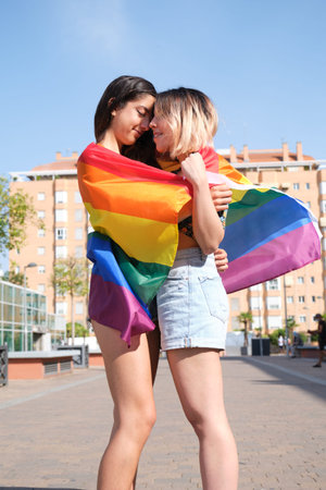 Young lesbian couple smiling and hugging wrapped in a rainbow flag. LGBT relationship.