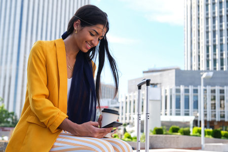 Young indian businesswoman wearing yellow suit jacket talking on the smartphone, holding a cup coffee and a suitcase sitting outdoor.の写真素材