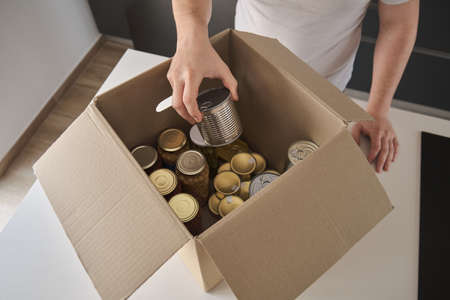 Unrecognizable young man filling donation box with food cans, legume jars and canned fish in kitchen.の写真素材