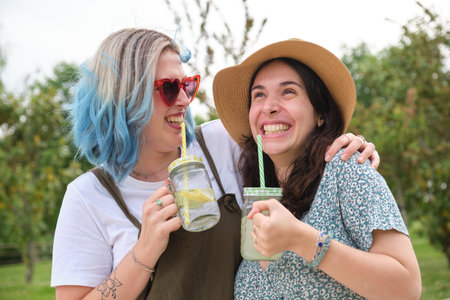 Two female happy friends drinking refreshing drinks, laughing, hugging and looking to each other in a park on a sunny summer day.の写真素材