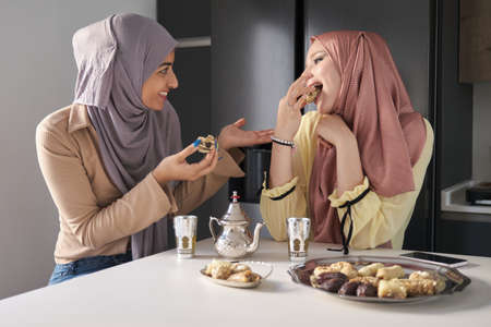 Two young muslim women talking, drinking tea and eating arabic sweets at the kitchen table.の写真素材