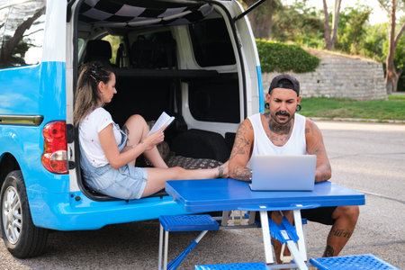 Young tattooed couple smiling, reading a book and working on a laptop sitting in a picnic table and...