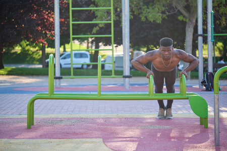 Young fit shirtless black man doing push-ups on parallel bars in a calisthenics park outdoors on...