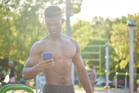 Portrait of a young fit shirtless black man using the smartphone in a calisthenics park outdoors....