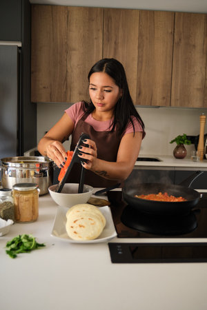 Young Venezuelan woman grating tomato to prepare the filling for the arepas at kitchen.の写真素材