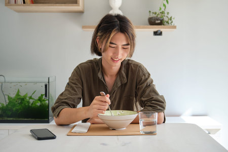 Smiley asian young man eating soup in the living room. Having lunch.