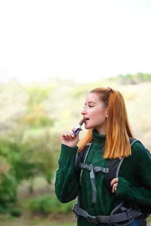 Hiker redhead woman eating a healthy no bake date bar, wearing a backpack. Trekking concept.