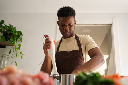 Young african man using a blender to prepare chicken mince in a kitchen.の写真素材