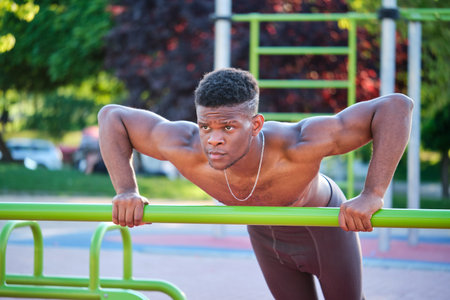 Young fit shirtless black man doing push-ups on a bar in a calisthenics park outdoors on sunny day....