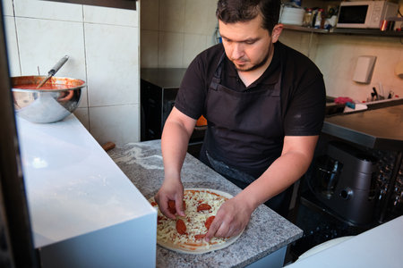 Latin man adding pepperoni to a raw pizza in a restaurant kitchen. Pizzeria.の写真素材