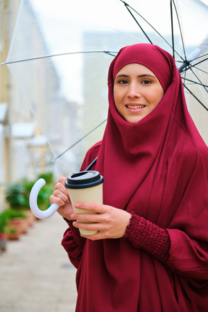 Portrait of muslim young woman in hijab smiling holding a coffee cup and an umbrella in the street and looking at camera.の写真素材