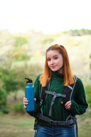 Smiley hiker redhead young woman holding canteen looking at camera in the mountain wearing a backpack. Trekking concept.の写真素材