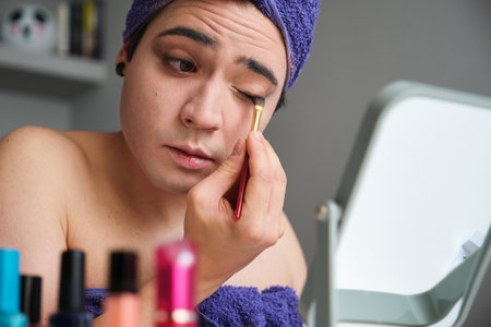 Young drag queen man applying eyeshadow after shower with a towel on his head.の写真素材