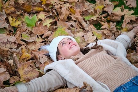 Preteen girl lying down on the ground covered in fallen leaves during autumn season. Happiness.