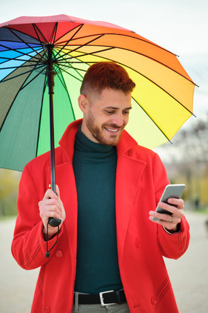 Latin young man with a red coat and a rainbow umbrella using a phone in the street in autumn.の写真素材