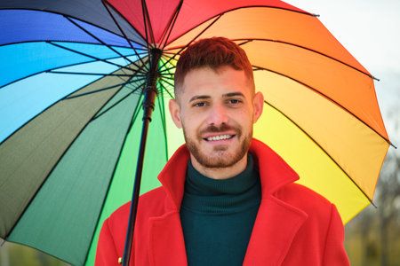 Portrait of latin young man with a red coat and a rainbow umbrella in the street in autumn.の写真素材