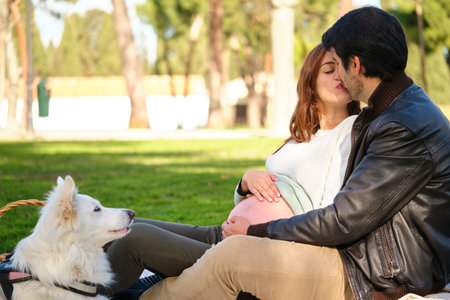Happy pregnant couple kissing on a picnic in a park with their dog.の写真素材