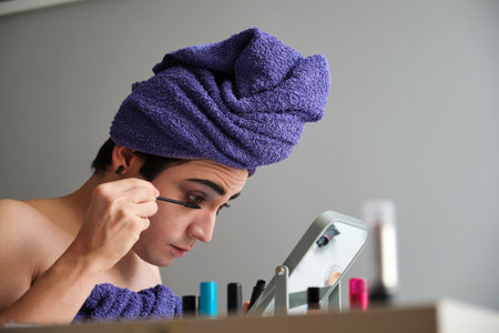 Young gender fluid person applying eye mascara after shower with a towel on his head.の写真素材