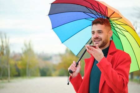 Latin young man with a red coat and a rainbow umbrella recording audio message on phone in the street in autumn.の写真素材