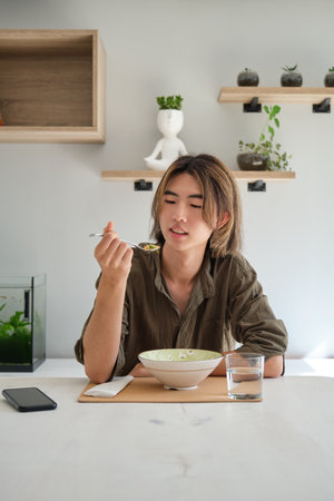 Smiling taiwanese young man eating soup in the living room. Having lunch.の写真素材