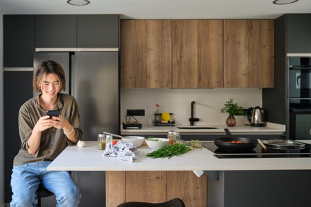 Smiling asian young man checking recipe on mobile phone at kitchen.