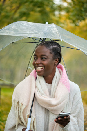 African woman with a smartphone and transparent umbrella smiling in autumn.の写真素材