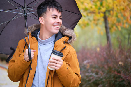 Smiling young man holding cup of coffee and umbrella in a rainy autumn day at the street.の写真素材