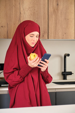 Muslim young woman in hijab drinking coffee and using smartphone in the kitchen at home.の写真素材