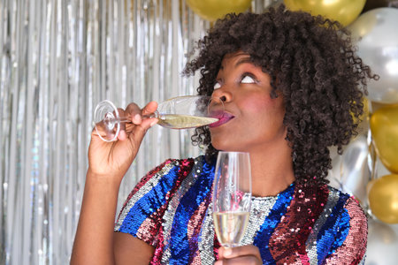 African woman drinking champagne holding two glasses celebrating New Year at a New Years Eve party.