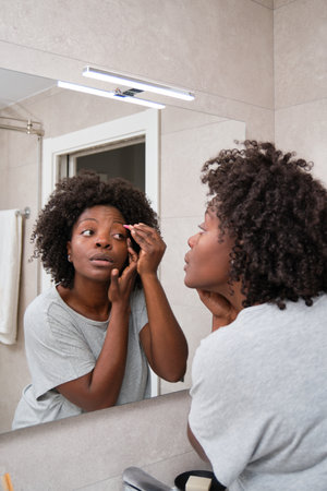 African young woman removing eyebrow hairs with tweezers in bathroom.の写真素材