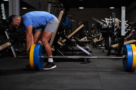 Strong Hispanic man putting weight plate on barbell before weightlifting at a gym.の写真素材
