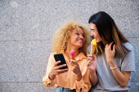 Two multiracial happy friends with ice creams using the smartphone in summer at street.