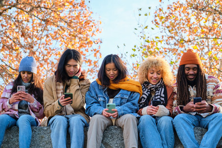 Group of multiracial young people using smart mobile phone outdoors in autumn. Happy university students watching cellphones sitting in college campus. Teenagers addicted to social media technology.の写真素材