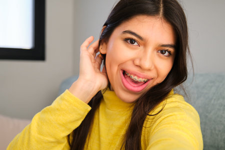 Happy Latin female teenager with braces taking a selfie at home.の写真素材