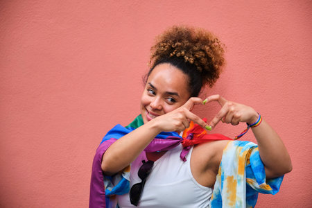 A woman with a rainbow flag as a cloak doing a heart shape with her hand. Celebrating Pride Day.