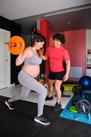 Pregnant woman exercising with a barbell with personal trainer's supervision at the gym.の写真素材