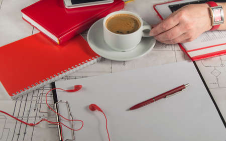 Female hand holding a Cup of coffee on the desktop with the drawings, red notebook and mobile phoneの写真素材